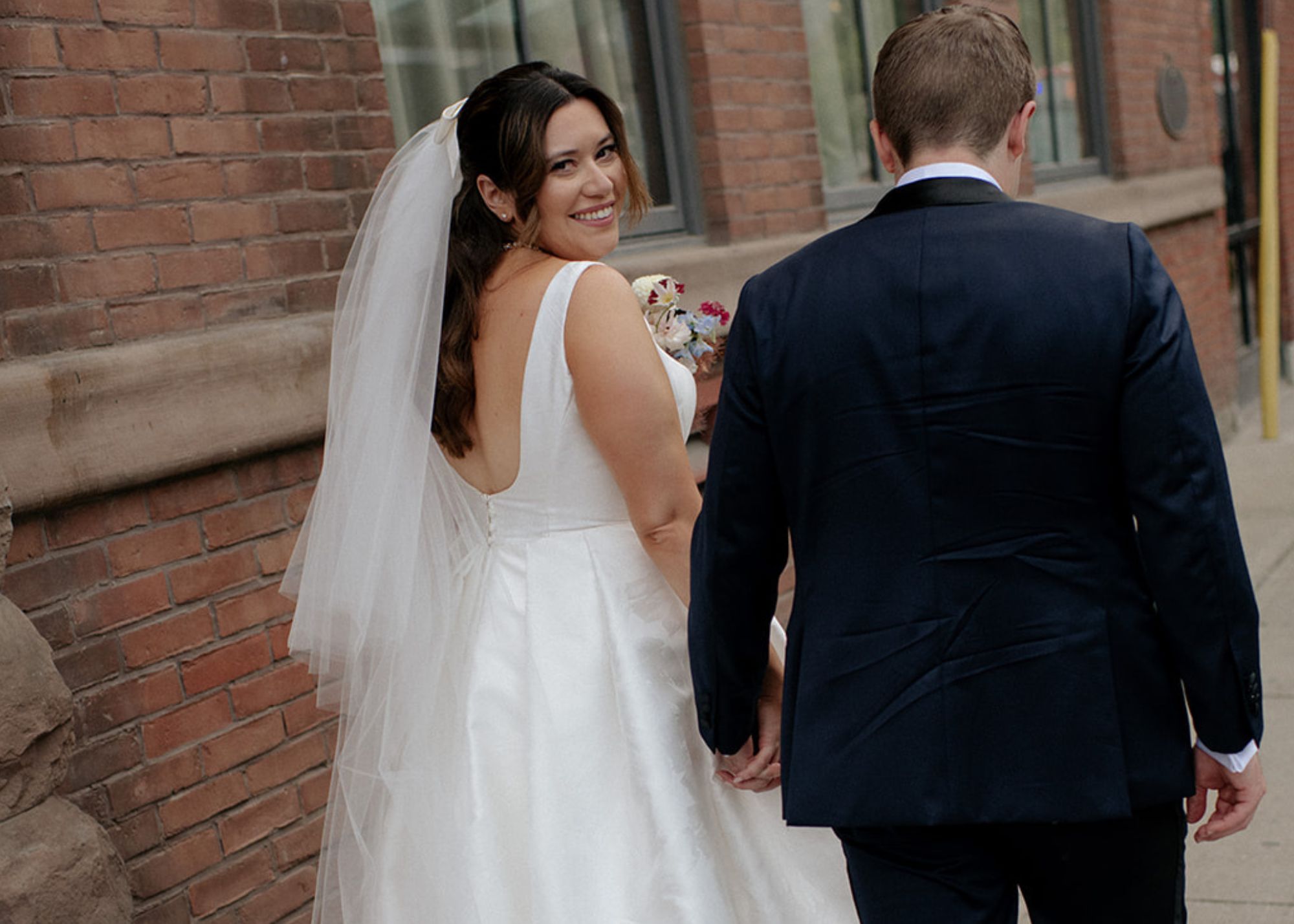 Newlyweds Savanna and Ian walk away from the camera. Savanna smiles over her shoulder and is wearing her custom square necked wedding dress