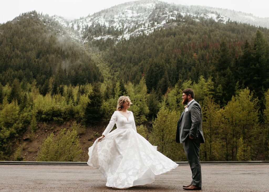 A bride twirls in her custom ball gown wedding dress with long sleeves as her groom watches on a misty day at Glacier National Park