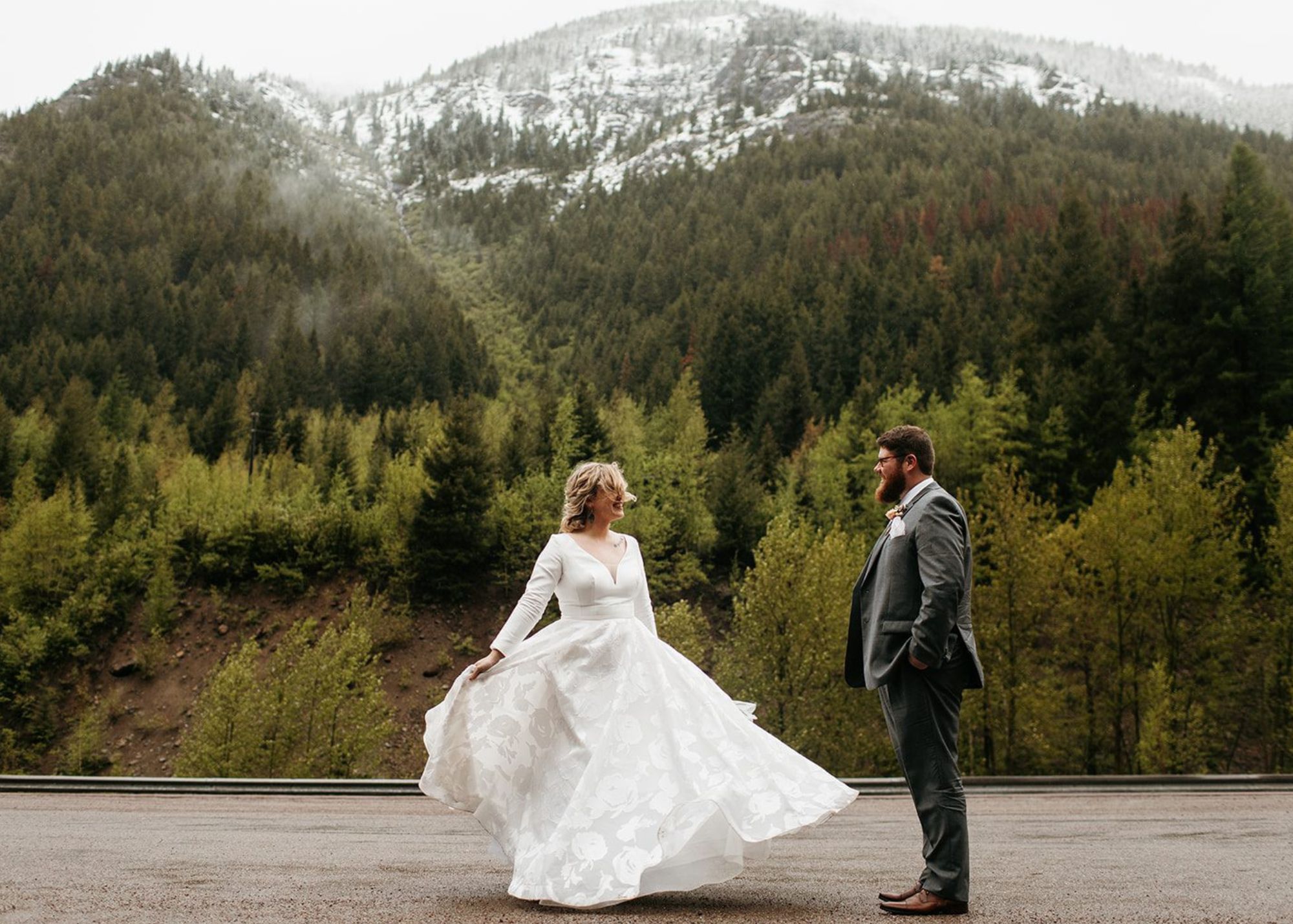 A bride twirls in her custom ball gown wedding dress with long sleeves as her groom watches on a misty day at Glacier National Park