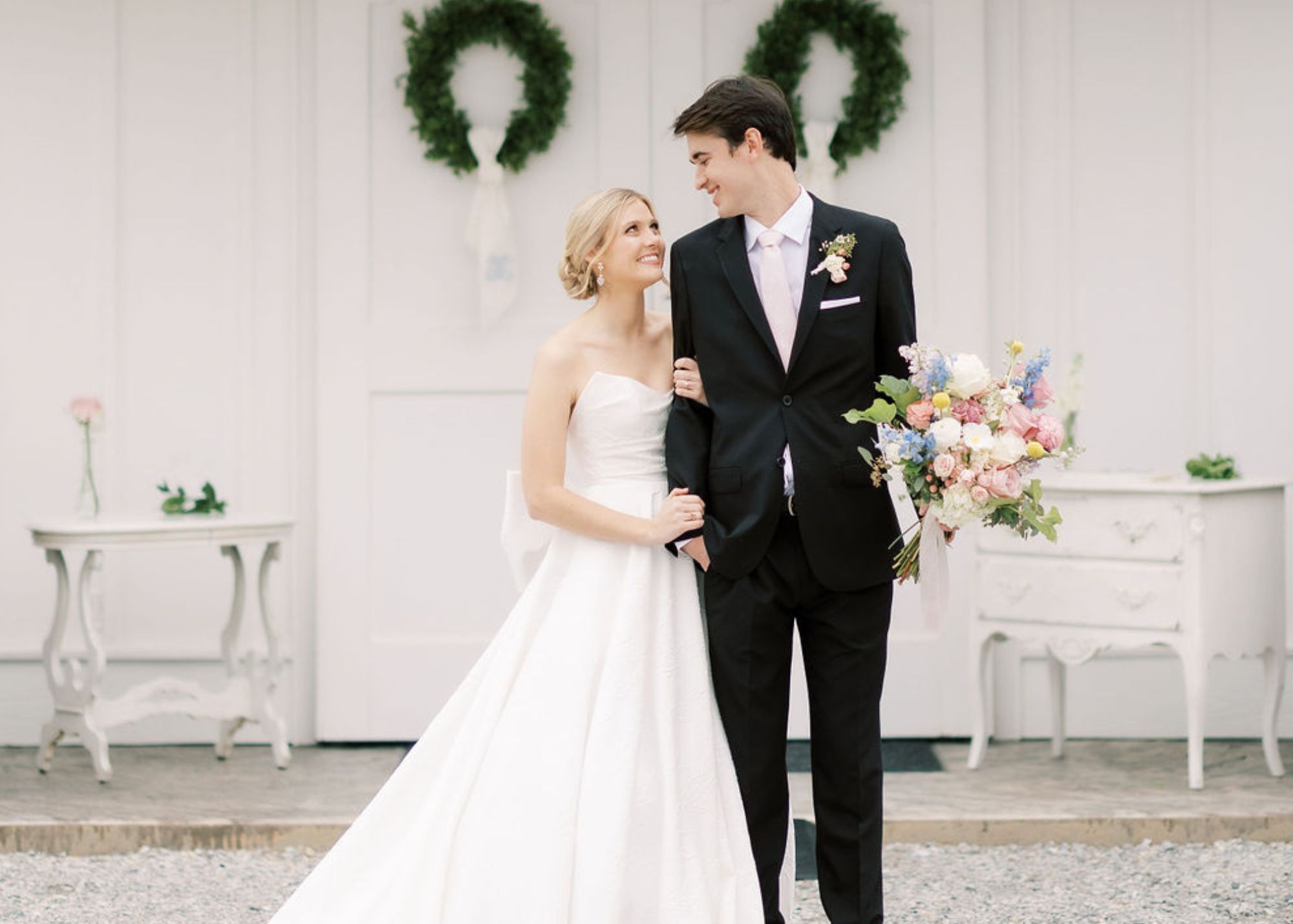 A bride and groom sand before white church doors with Christmas wreaths. The bride wears an a-line strapless wedding gown.