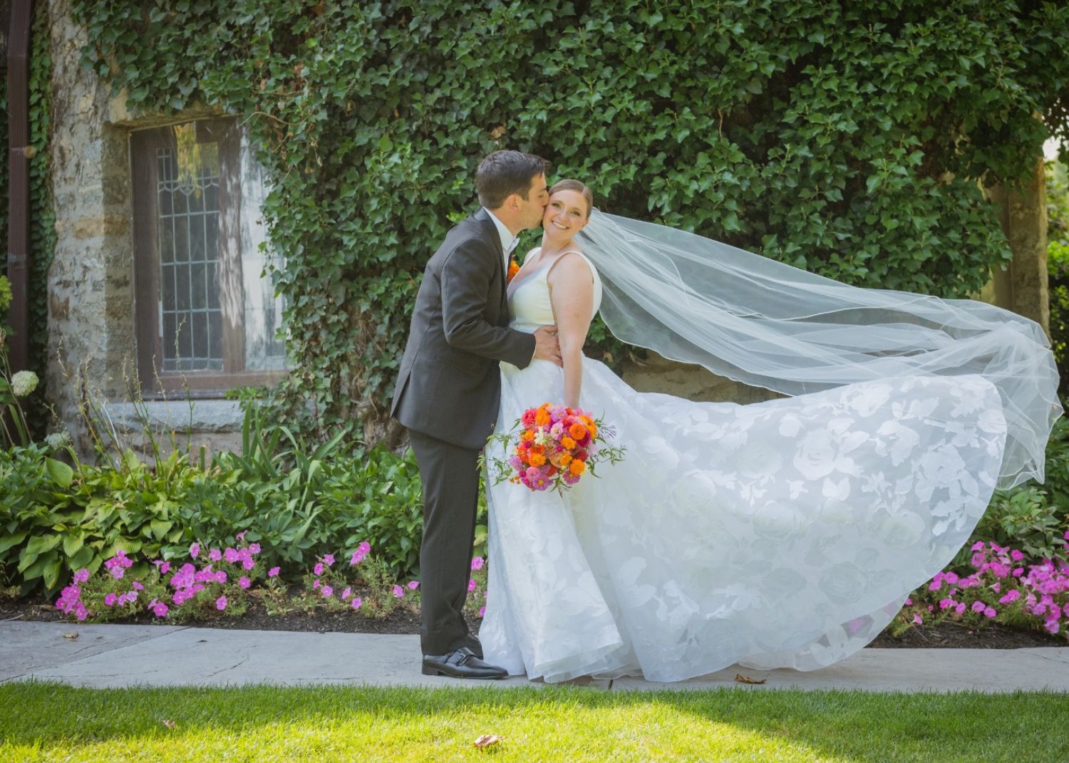 Real bride Lauren beams as her groom ikisses her and her flowing A-line wedding dress and veil float behind her in the summer sunshine