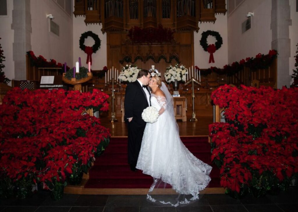 Sara and Parker stand at the later amidst poinsettias and evergreens. Sea is wearing a lace ball gown wedding dress and wrap by Lea-Ann Belter Bridal