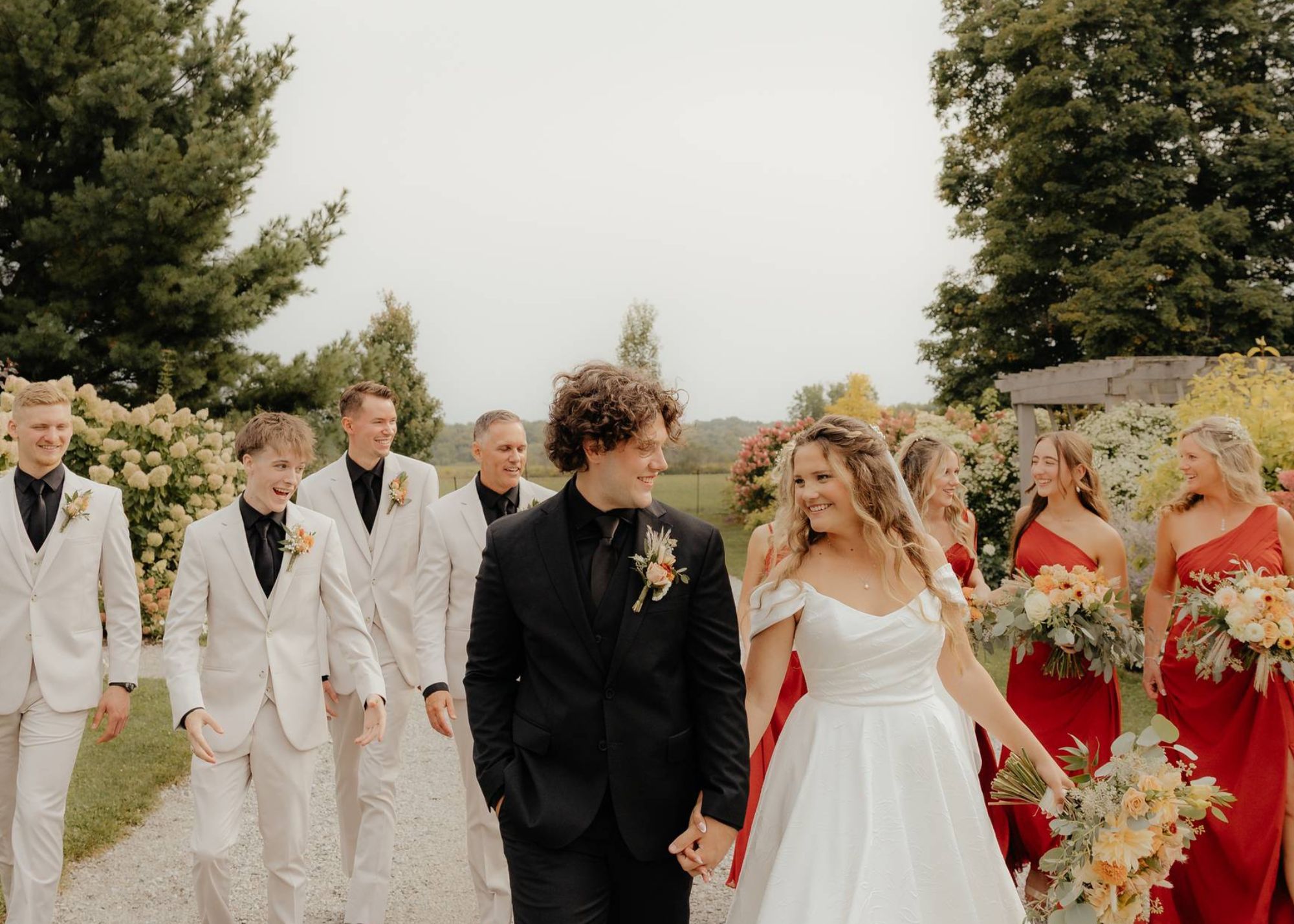 a bride and groom walk toward the camera in a meadow, their wedding party behind them. The bride wears a custom sized Astrid & Mercedes Rosebud gown with off-the-shoulder sleeves. The groom wears a black shirt and black suit.