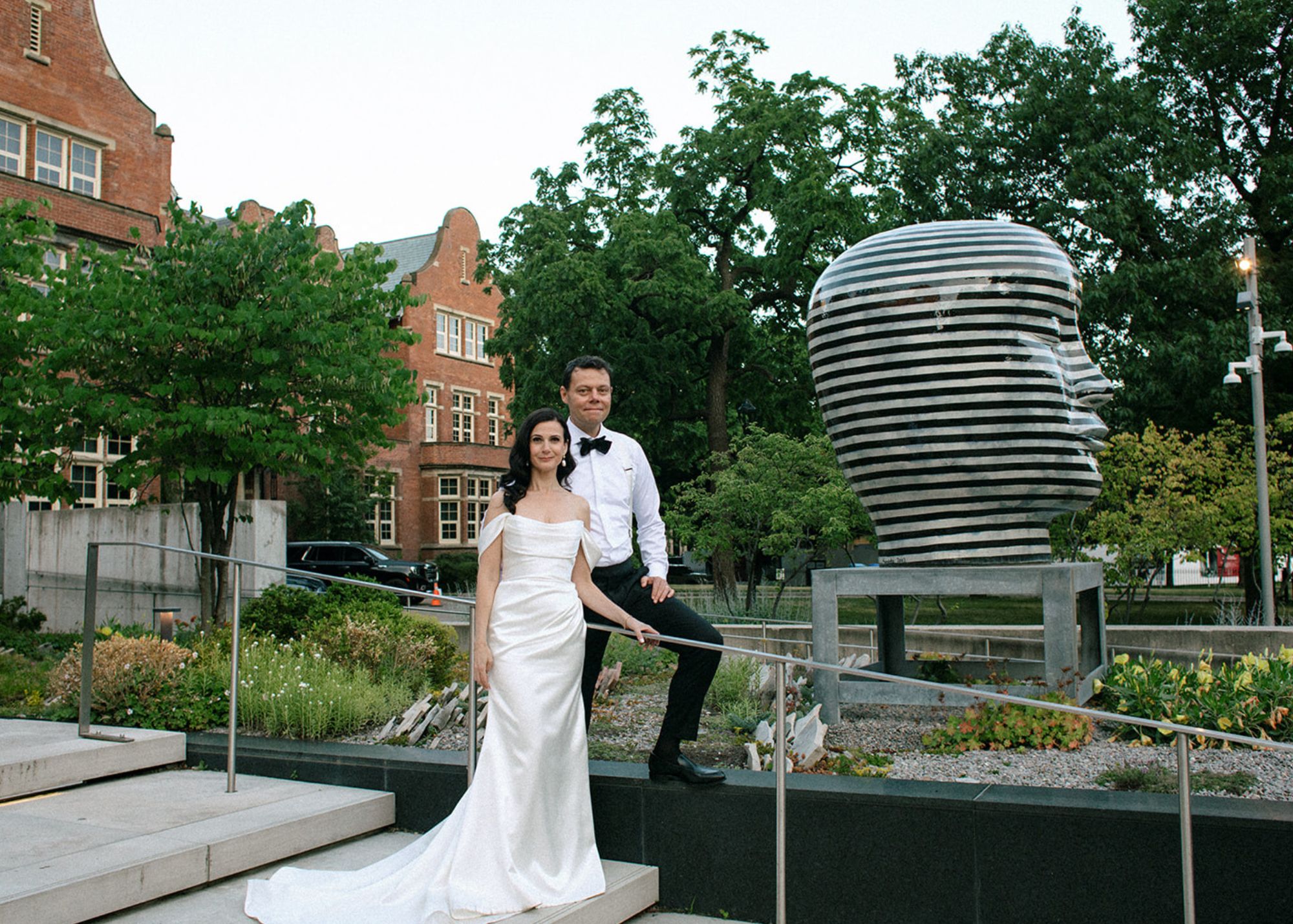 A bride and groom stand outdoors on the steps of Toronto's Gardiner Museum before a modern sculpture. The bride wears a sleek wedding dress by Lea-Ann Belter Bridal, and the groom wears a tux without the jacket.