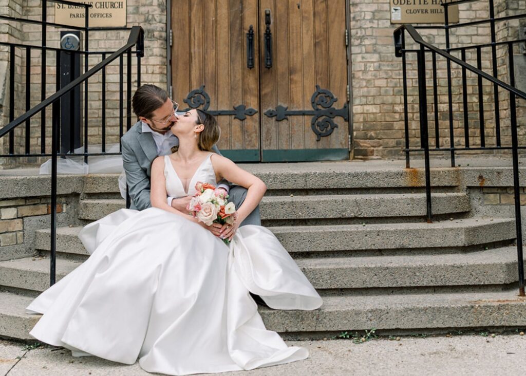 A rideand groom sit on the church steps, sharing a passionate kiss. The bride's v-neck wedding gown with full skirt.