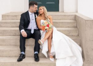 A bride and groom sit on steps, holding one another. The bride wears Liliana, a strapless ballgown with a thigh-high slit