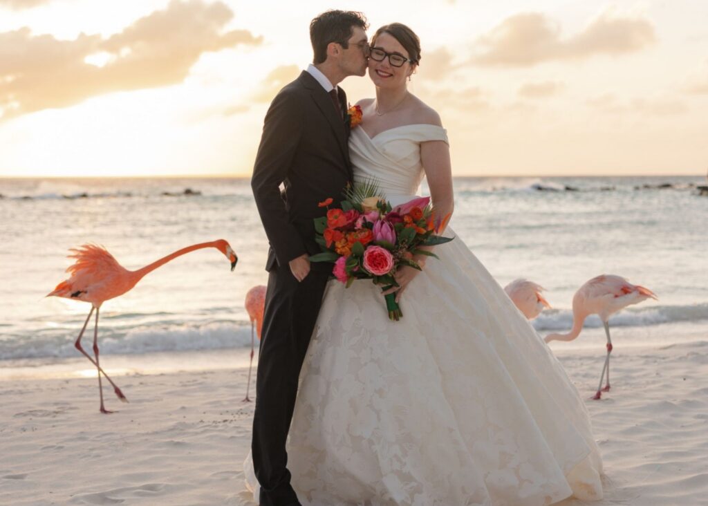 A groom kisses his smiling bride on the cheek on a beach at sunset in Aruba as flamingos walk around them. The bride wears glasses, and a custom off-the-shoulder ball gown wedding dress.
