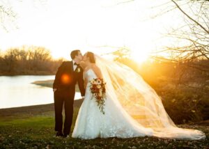 A bride and groom kiss before a lake during golden hour. The bride's cathedral veil and ball gown flutter behind her in the breeze.