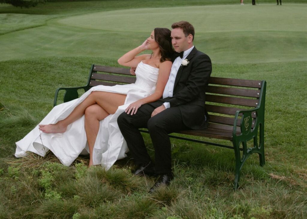 a bride in a corset bodice ball gown wedding dress with a high slit sits on a bench in a green park beside her groom.