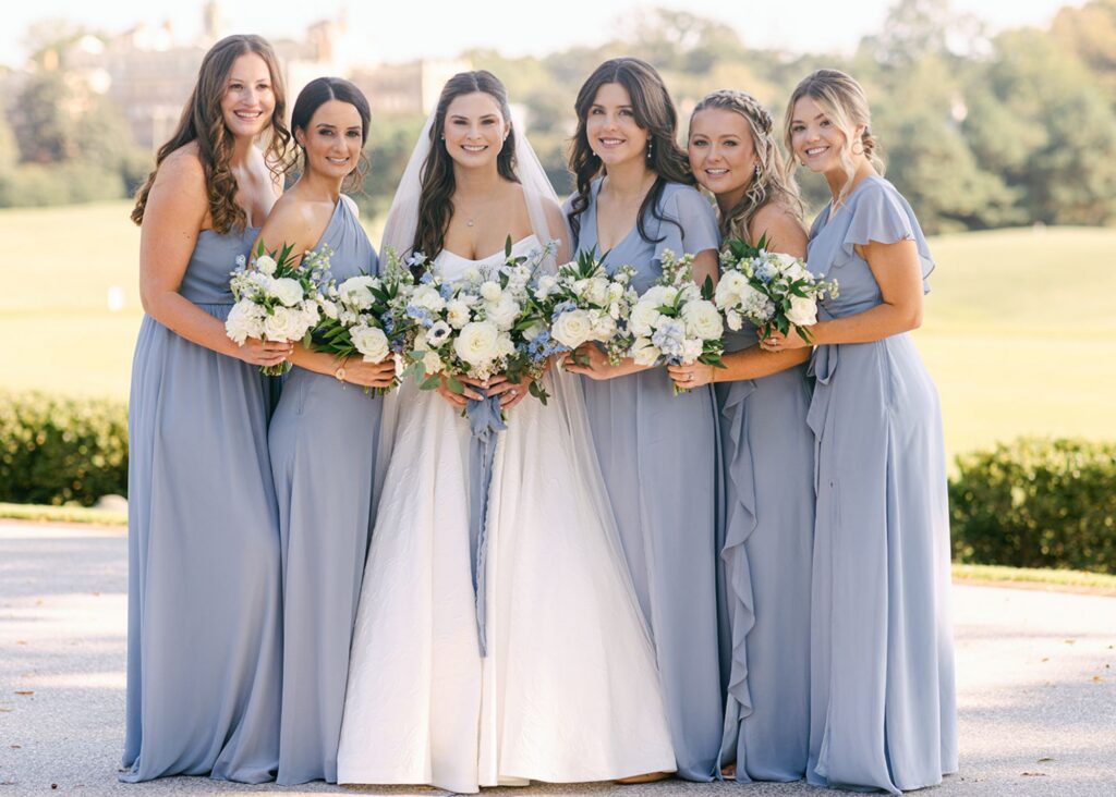 a bride and her bridesmaids stand together in the sun. The bride wears an A-line wedding dress, and her bridesmaids wear a lovely hydrangea blue.