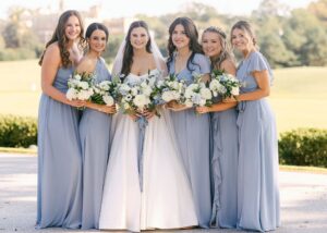 a bride and her bridesmaids stand together in the sun. The bride wears an A-line wedding dress, and her bridesmaids wear a lovely hydrangea blue.