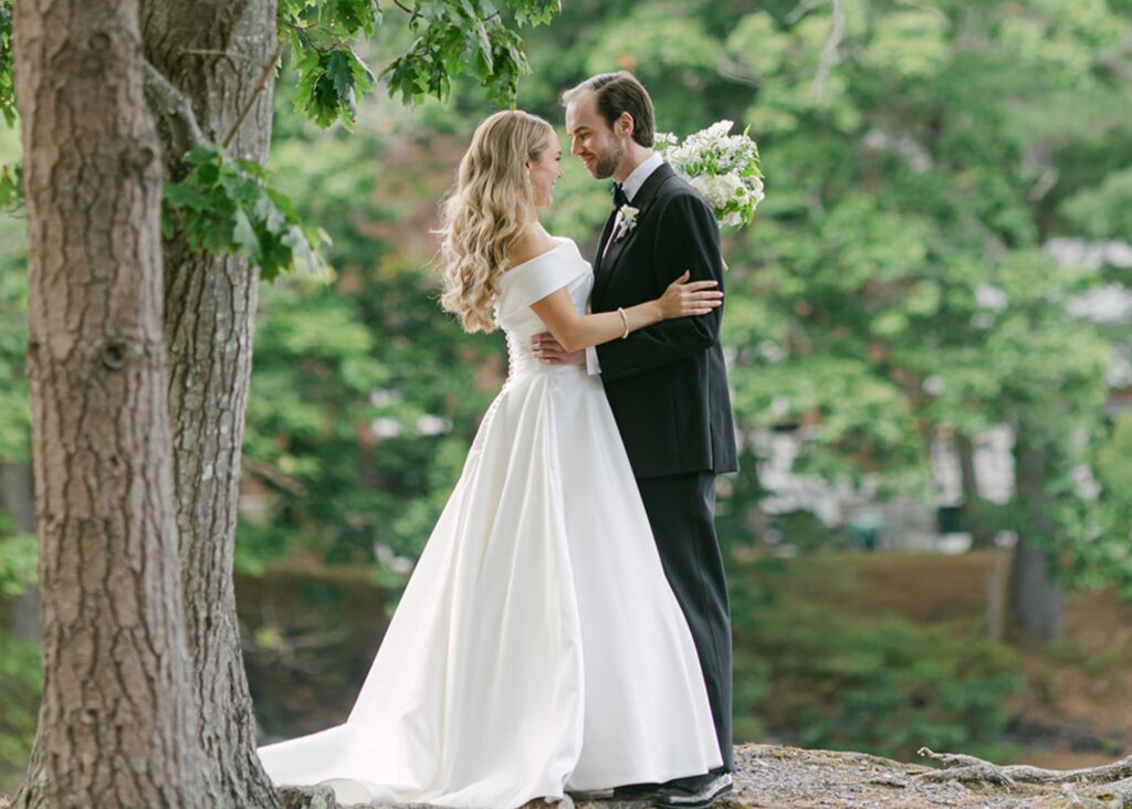 A bride and groom stand in one another's arms beneath a tree in the Maine Woods. The bride wears her customized Hannah ball gown wedding dress by Lea-Ann Belter Toronto.