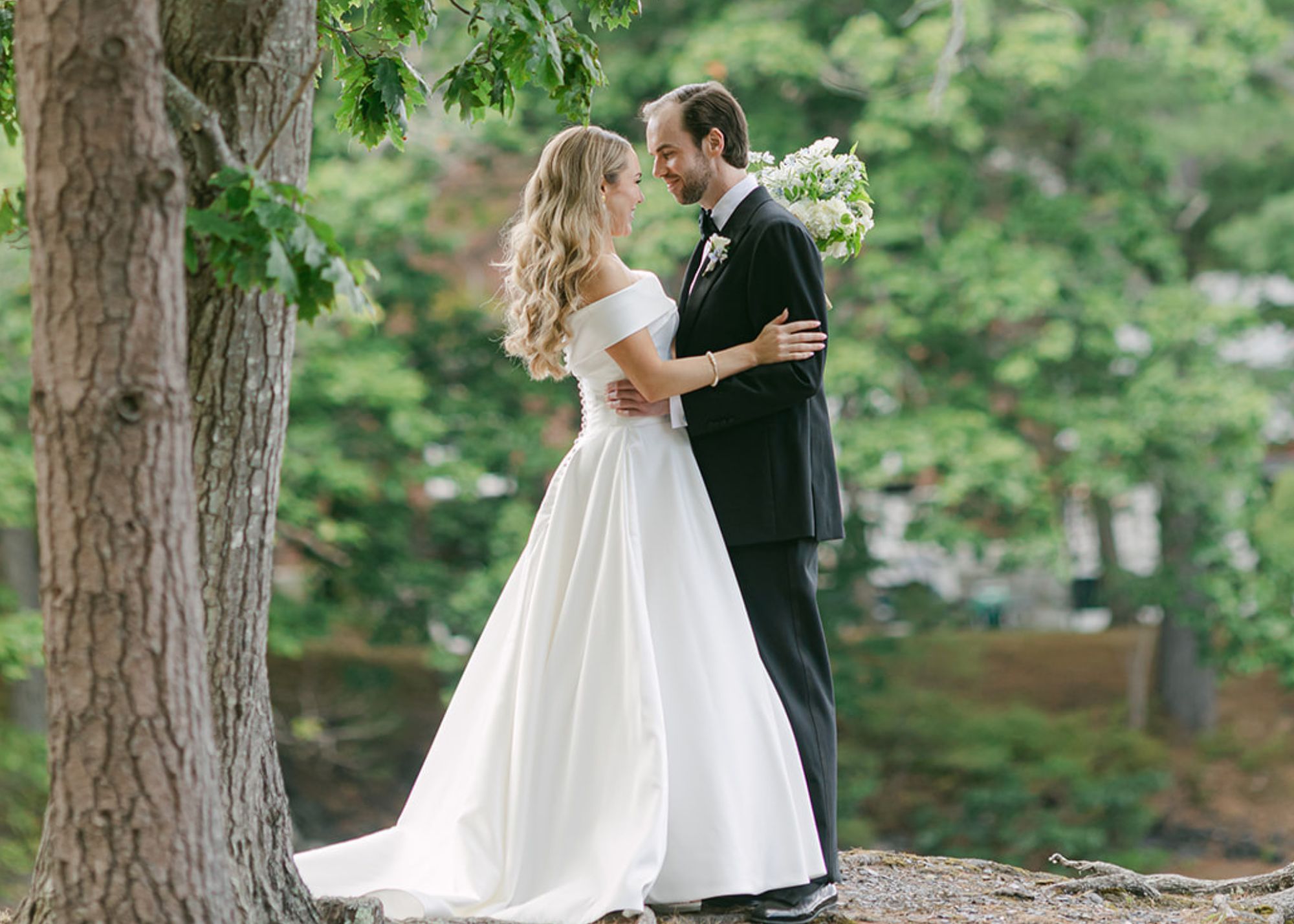 A bride and groom stand in one another's arms beneath a tree in the Maine Woods. The bride wears her customized Hannah ball gown wedding dress by Lea-Ann Belter Toronto.
