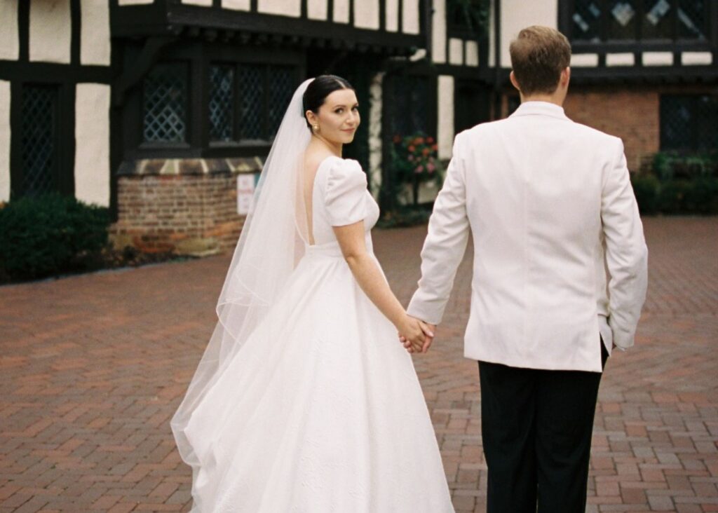 A bride and groom walk away from the camera, the bride looks over her shoulder. The bride is wearing a puff sleeved wedding ball gown and cathedral veil, the groom a white dinner jacket.