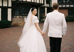 A bride and groom walk away from the camera, the bride looks over her shoulder. The bride is wearing a puff sleeved wedding ball gown and cathedral veil, the groom a white dinner jacket.