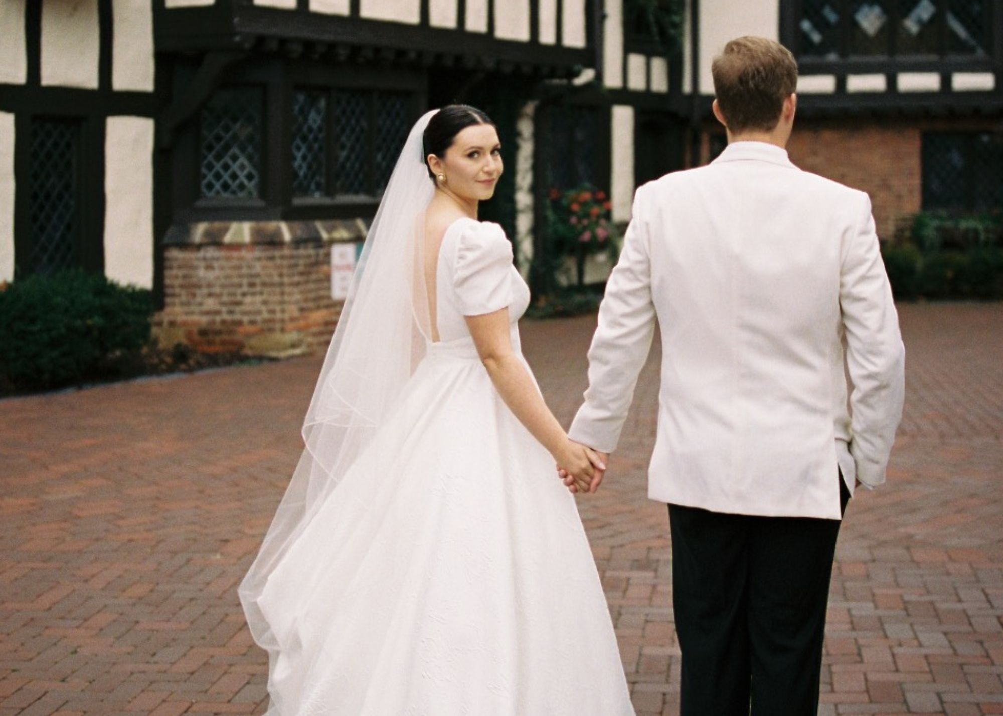 A bride and groom walk away from the camera, the bride looks over her shoulder. The bride is wearing a puff sleeved wedding ball gown and cathedral veil, the groom a white dinner jacket.