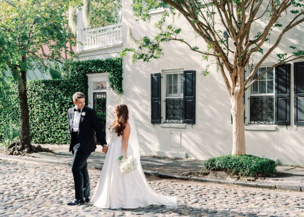 A bride and groom walk hand in hand on a cobblestoned street in Charleston. The bride wears a classic a-line wedding dress and a flowing veil. The groom wears a tuxedo.