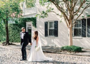 A bride and groom walk hand in hand on a cobblestoned street in Charleston. The bride wears a classic a-line wedding dress and a flowing veil. The groom wears a tuxedo.