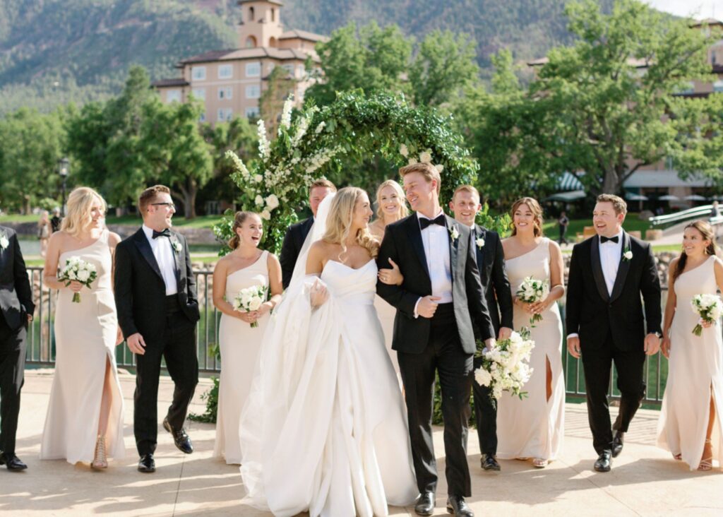 A bride and groom walk in the sunshine smiling at one another as their wedding party follows. The bride is weaing a ball gown wedding dress and a cathedral veil, and the groom wears a tux