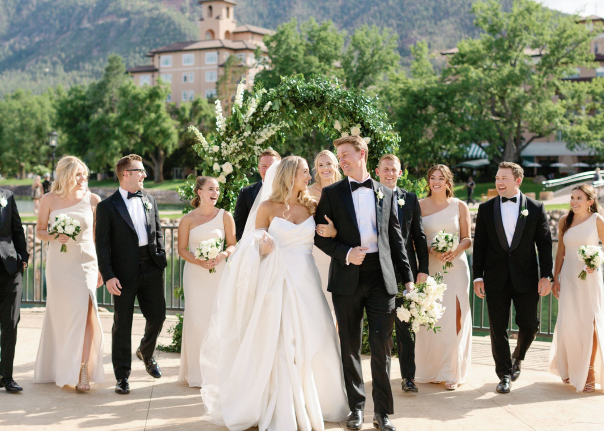 A bride and groom walk in the sunshine smiling at one another as their wedding party follows. The bride is weaing a ball gown wedding dress and a cathedral veil, and the groom wears a tux