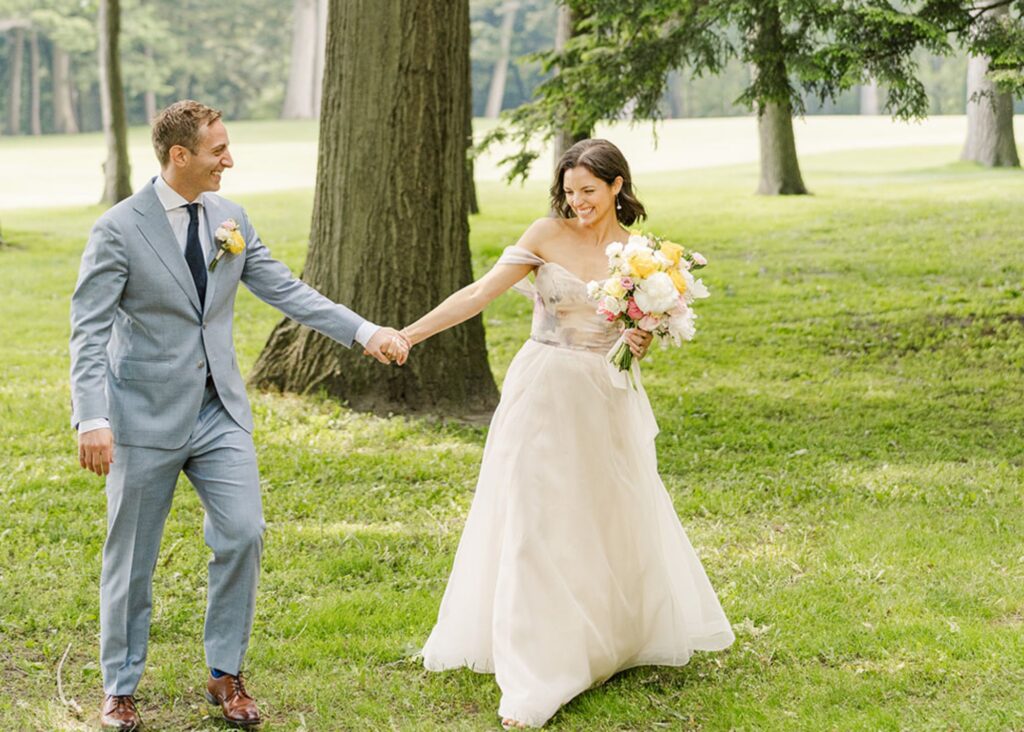A bride and groom walk hand in hand laughing in a wood. The bride wears her custom a-line floral print wedding gown and carries her yellow and white bouquet.
