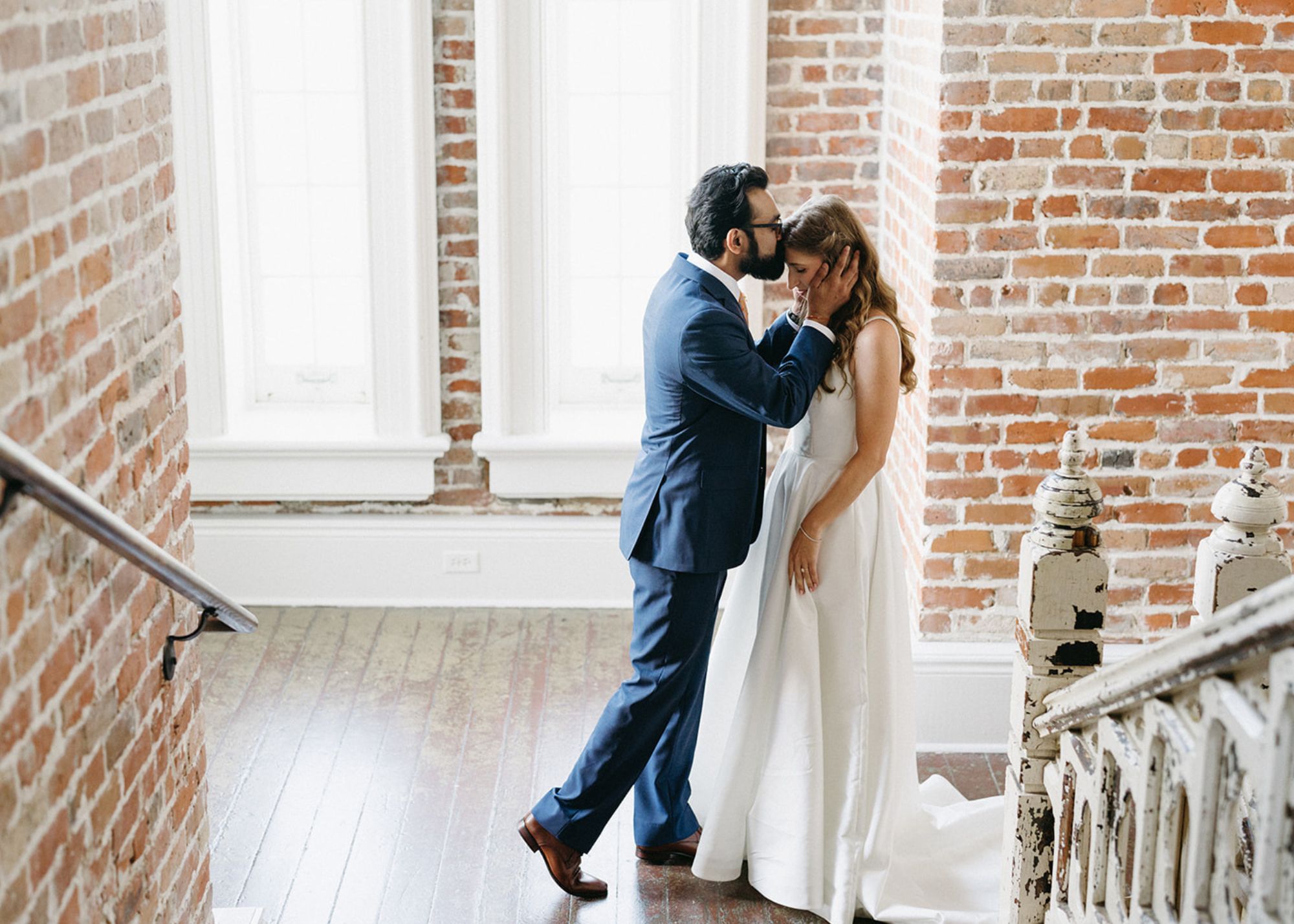 a groom sweetly kisses his bride's forehead in a sunlight drenched stairwell. the bride wears a square neck ball gown wedding dress