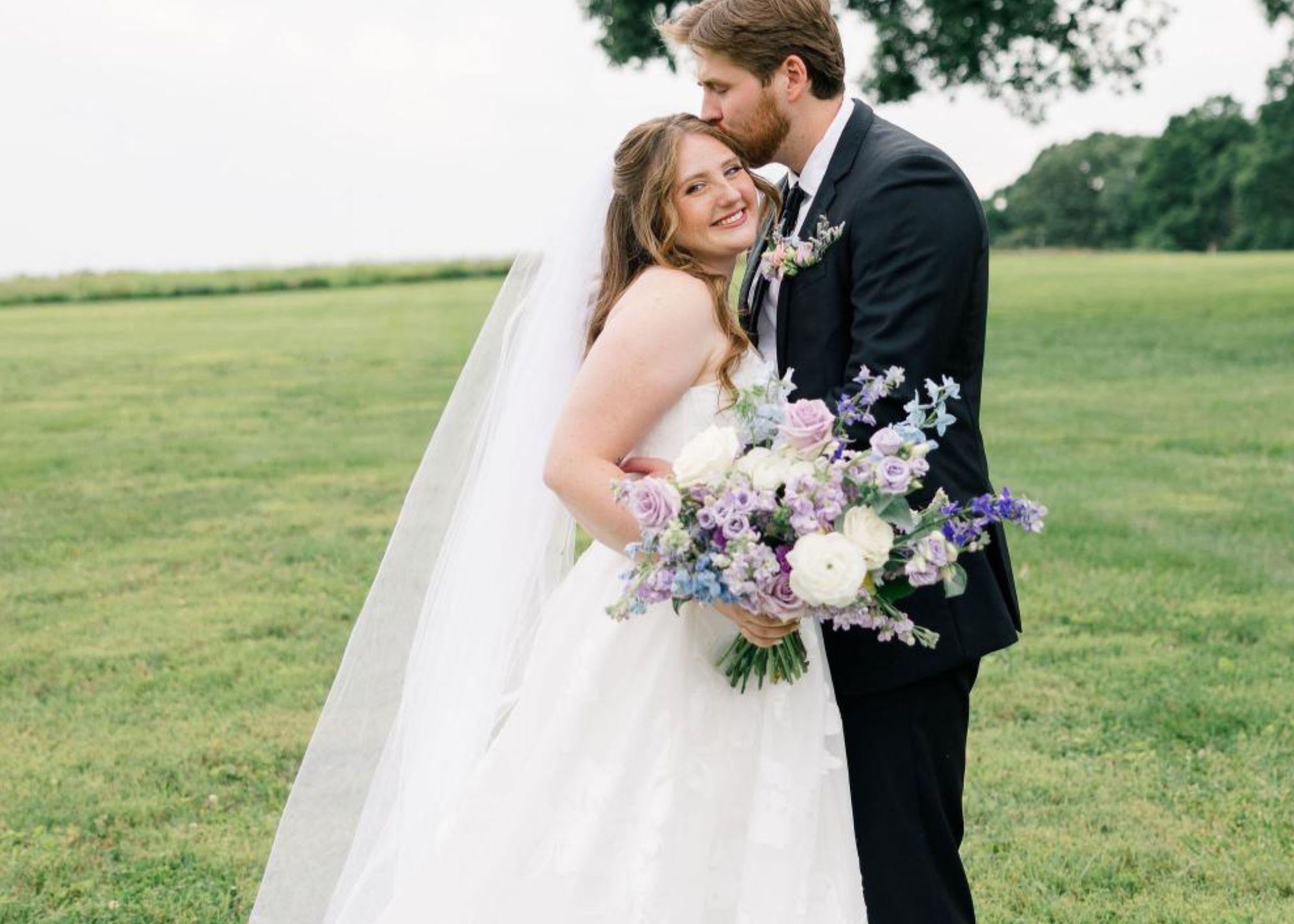 a bride is held he her groom's arms and smiles at the camera as he kisses her head. She's wearing a floral organza ball gown wedding dress by Lea-Ann Belter Bridal
