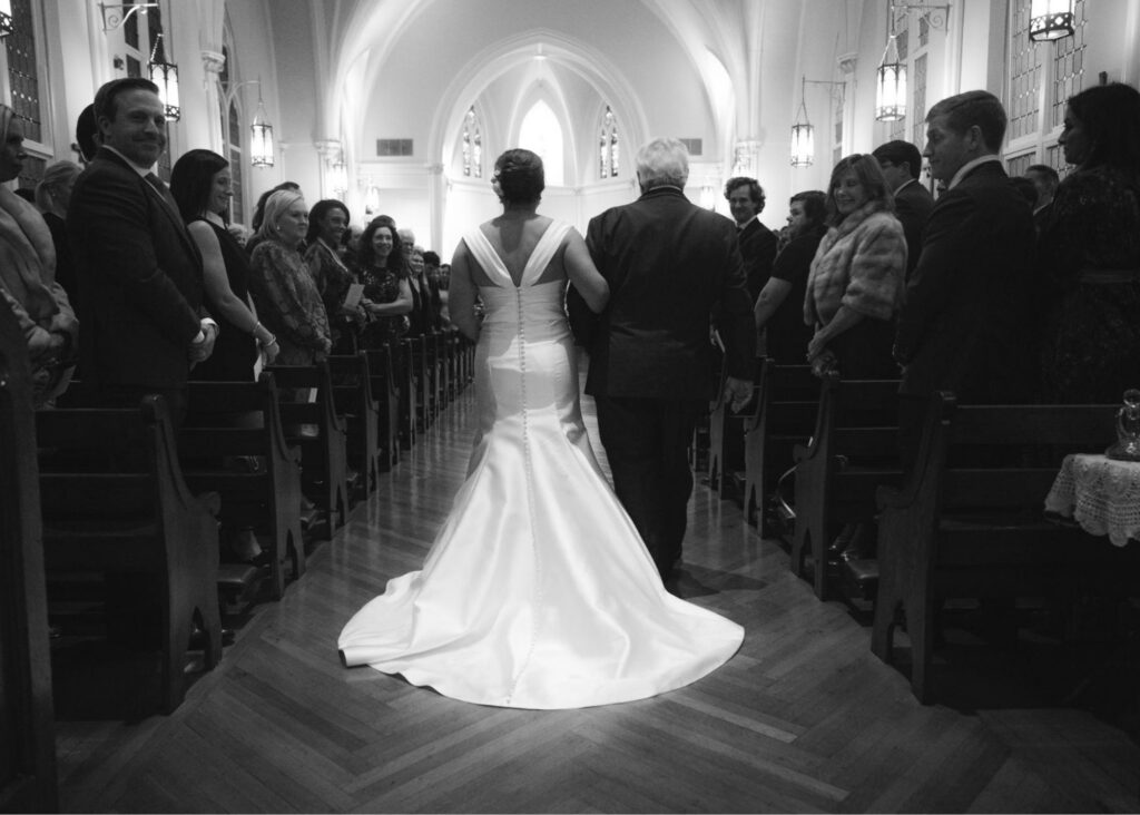 A black and white photo of a bride in a fit and flare wedding dress walking arm and arm with her father down the aisle from behind.