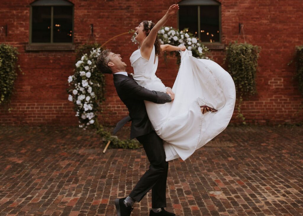 A jubilant bride and groom celebrate before a crescent floral arbor. The bride wears a sleek wedding dress and as the groom lifts her she kicks her feet, making the dress's silk fabric billow.