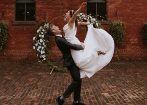 A jubilant bride and groom celebrate before a crescent floral arbor. The bride wears a sleek wedding dress and as the groom lifts her she kicks her feet, making the dress's silk fabric billow.