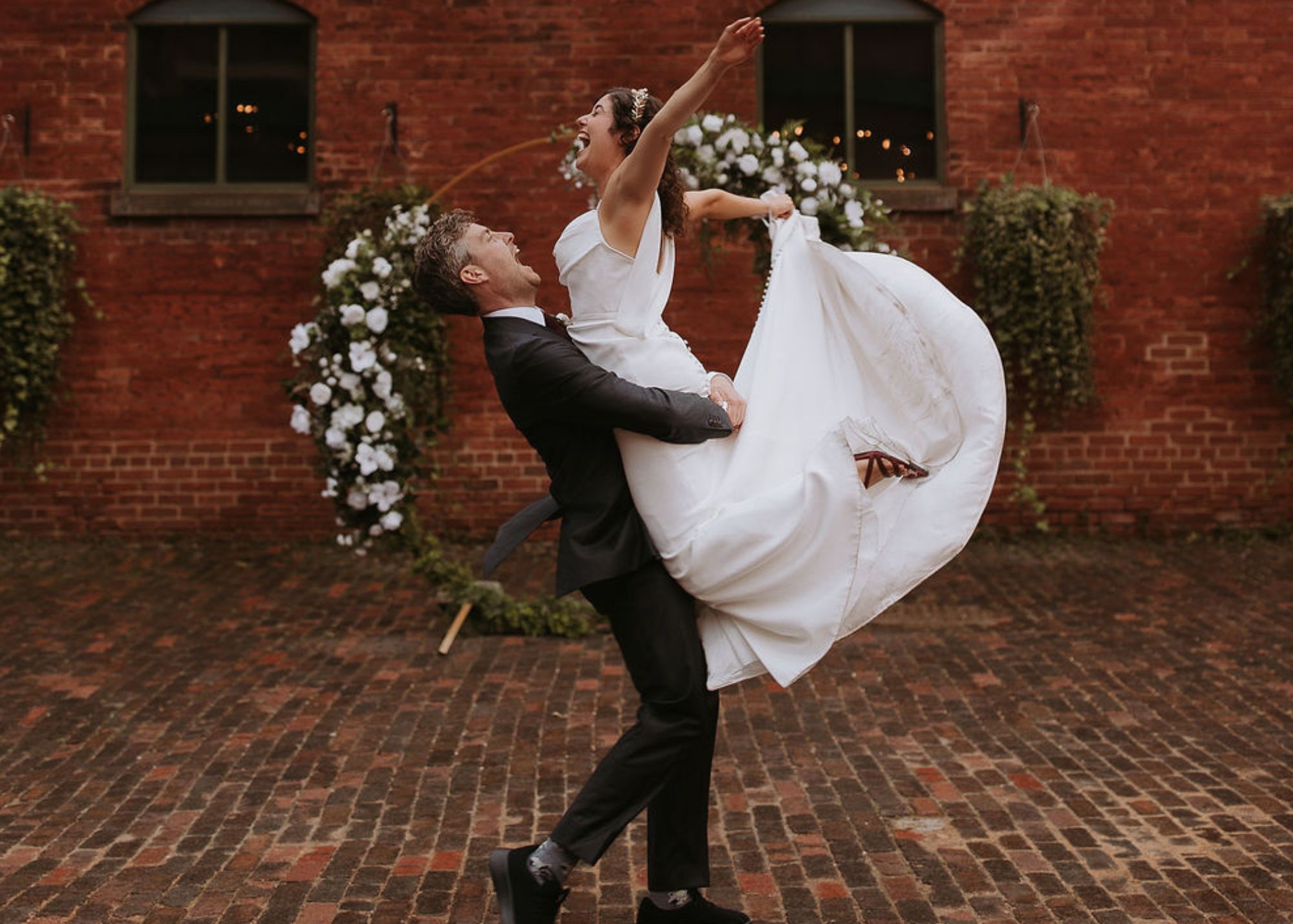 A jubilant bride and groom celebrate before a crescent floral arbor. The bride wears a sleek wedding dress and as the groom lifts her she kicks her feet, making the dress's silk fabric billow.