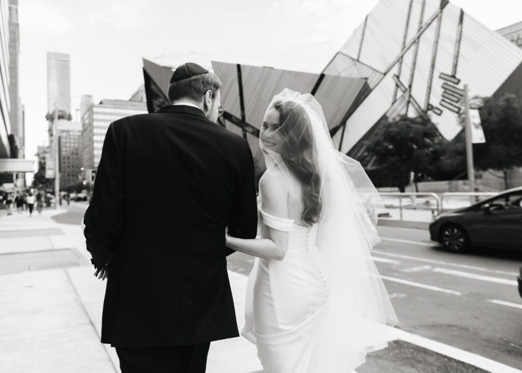 A black and white photo of a bride and groom walking away. The bride is wearing a sleek wedding dress and cathedral veil and looks over her shoulder