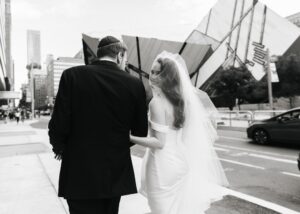 A black and white photo of a bride and groom walking away. The bride is wearing a sleek wedding dress and cathedral veil and looks over her shoulder