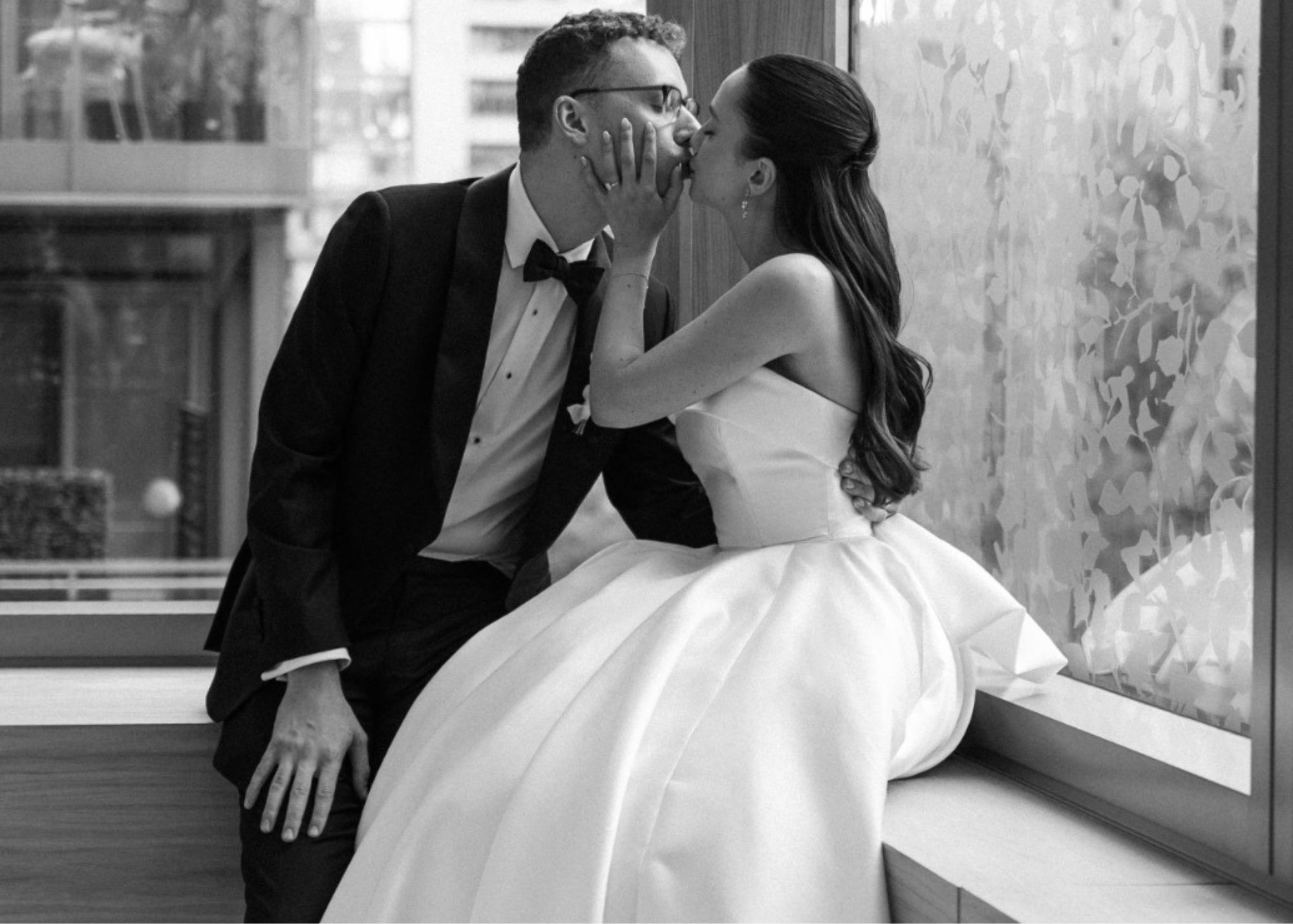 A black and white photo o f bride and groom seated beside one another, kissing. The. bride wears a strapless ball gown wedding dress by Lea-Ann Belter, Toronto