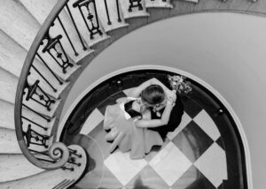 ⁠a black and white overhead shot of a bride and groom kissing. The bride wears a bow in her hair and a custom tea-length wedding dress inspired by Audrey Hepburn’s film, Funny Face