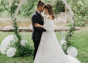 A bride and groom stand before a circular floral backgrop in a garden wedding ceremony, nose to nose. The brides wears a floral organza ball gown wedding dress, the groom a black tux.