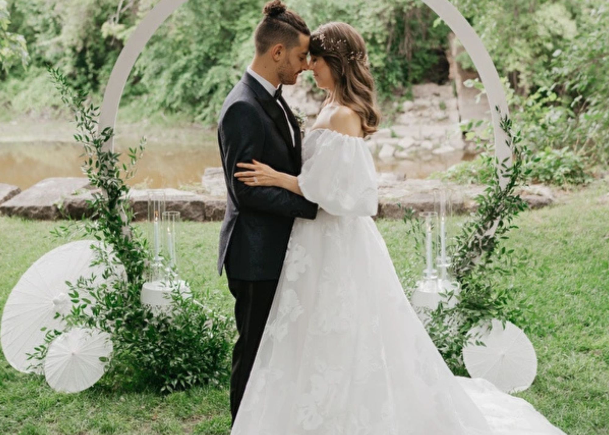 A bride and groom stand before a circular floral backgrop in a garden wedding ceremony, nose to nose. The brides wears a floral organza ball gown wedding dress, the groom a black tux.