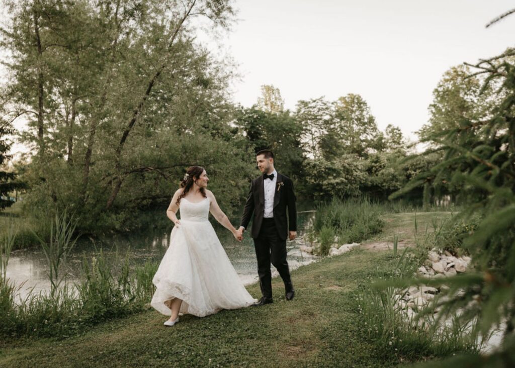 A bride and groom walk hand in hand along a wooded stream. The bride wears a custom wedding dress by Lea-Ann Belter, the groom a tux