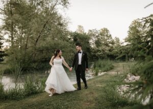 A bride and groom walk hand in hand along a wooded stream. The bride wears a custom wedding dress by Lea-Ann Belter, the groom a tux