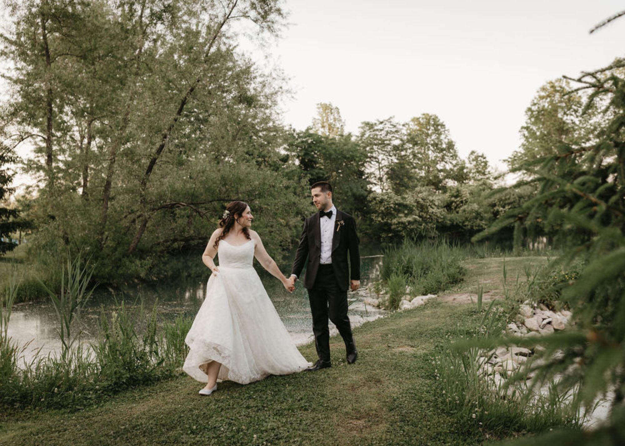 A bride and groom walk hand in hand along a wooded stream. The bride wears a custom wedding dress by Lea-Ann Belter, the groom a tux