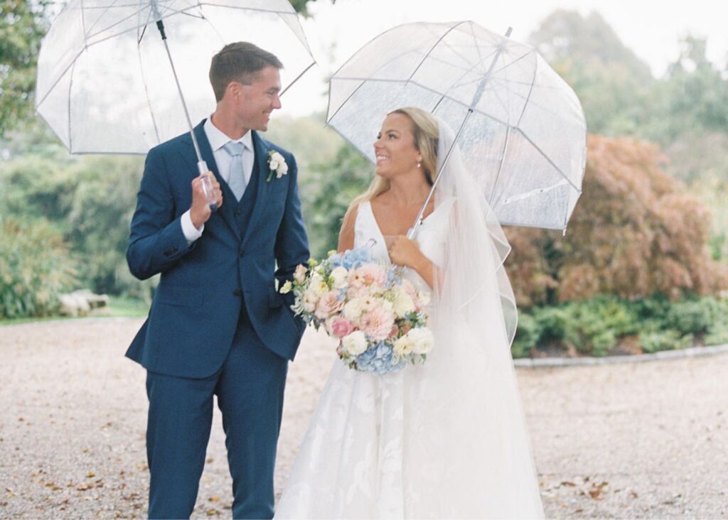 A bride and groom smile up at one another from underneath clear umbrellas> the groom wears a navy suit, the bride a v-neck wedding dress with organza A-line skirt