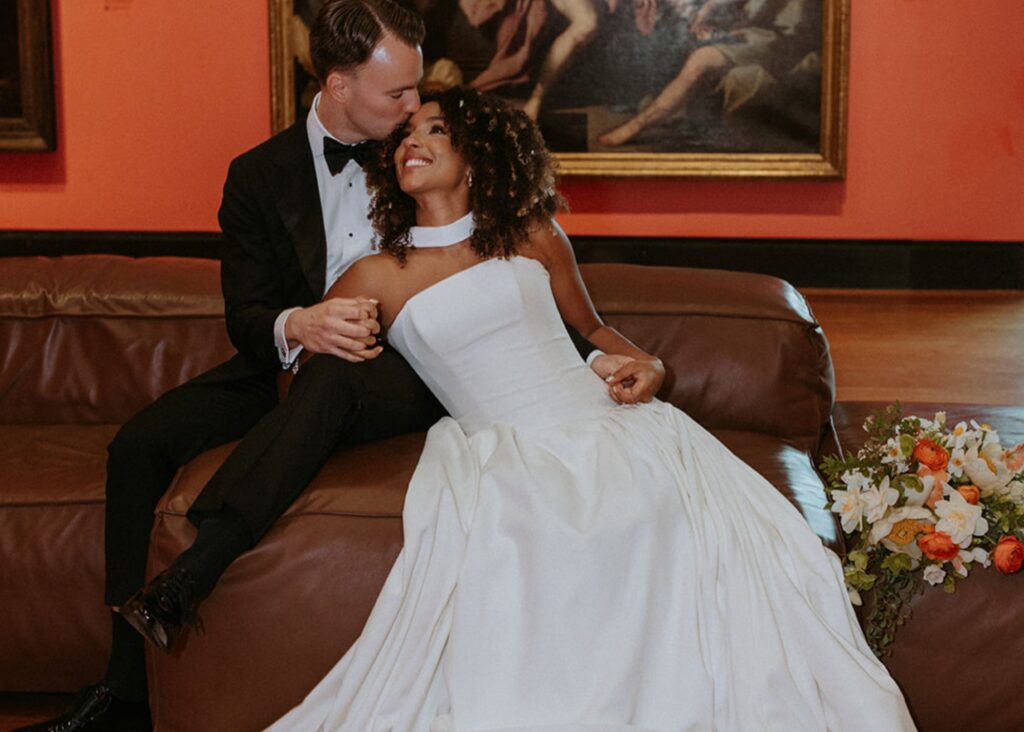 A bride wearing a modern drop waist ball gown of fluid crepe leans into her groom within the dramatic halls of the Art Gallery of Ontario.