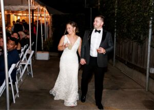 A bride and groom walk hand in hand beside their reception tent. The bride wears a lace fitted wedding dress, the groom a tux