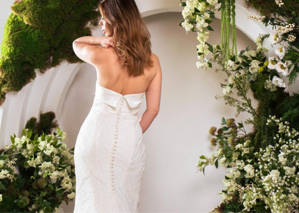 Bride wearing Chloe by Lea-Ann Belter Bridal, a strapless lace fit-and-flare wedding dress with bow back detail and button-traced train, surrounded by lush florals