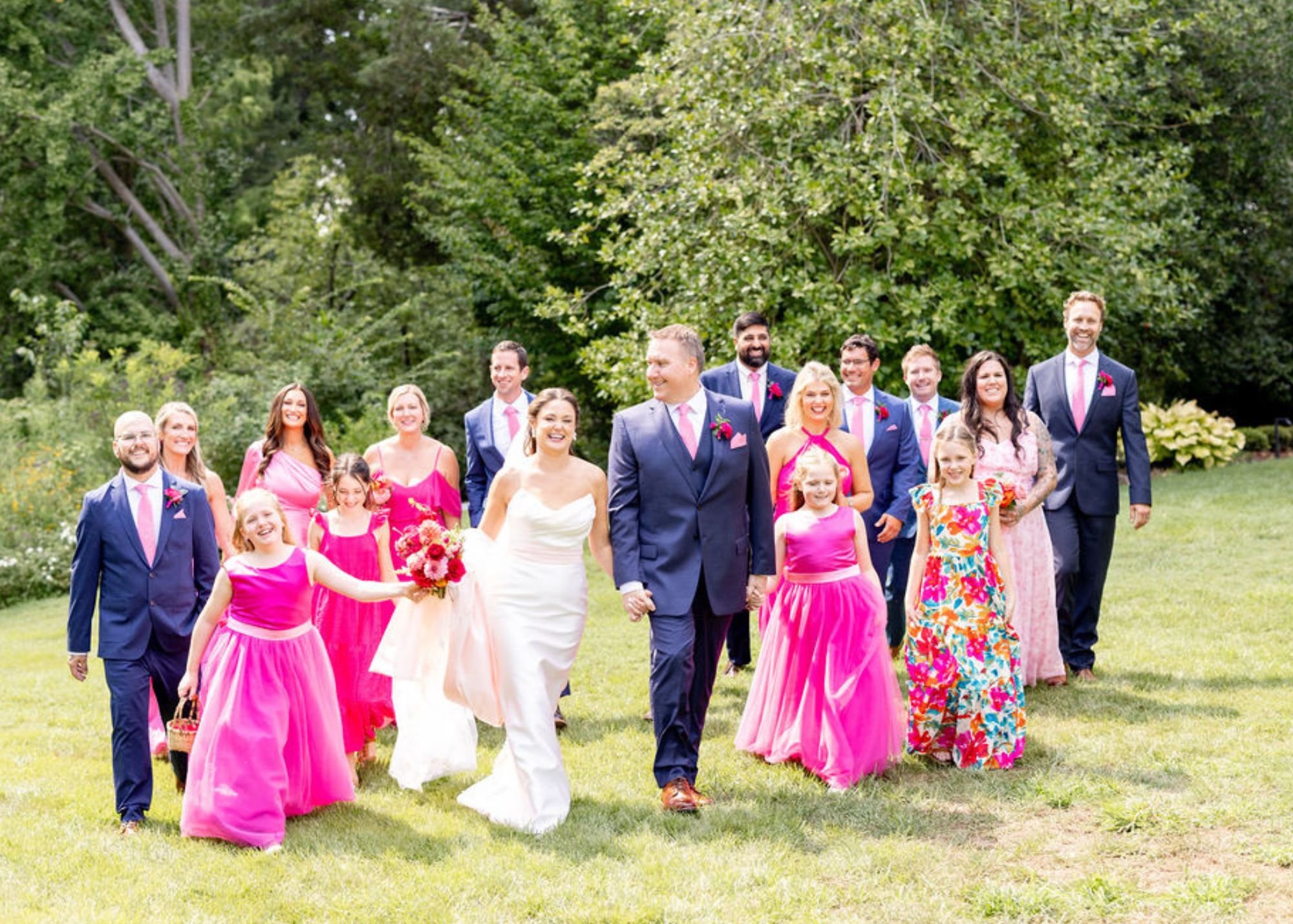 a bride and groom walk across a sunny lawn with their wedding party. The bride wears a sleek column bridal dress with a pink sash, and the bridesmaids wear hot pink.