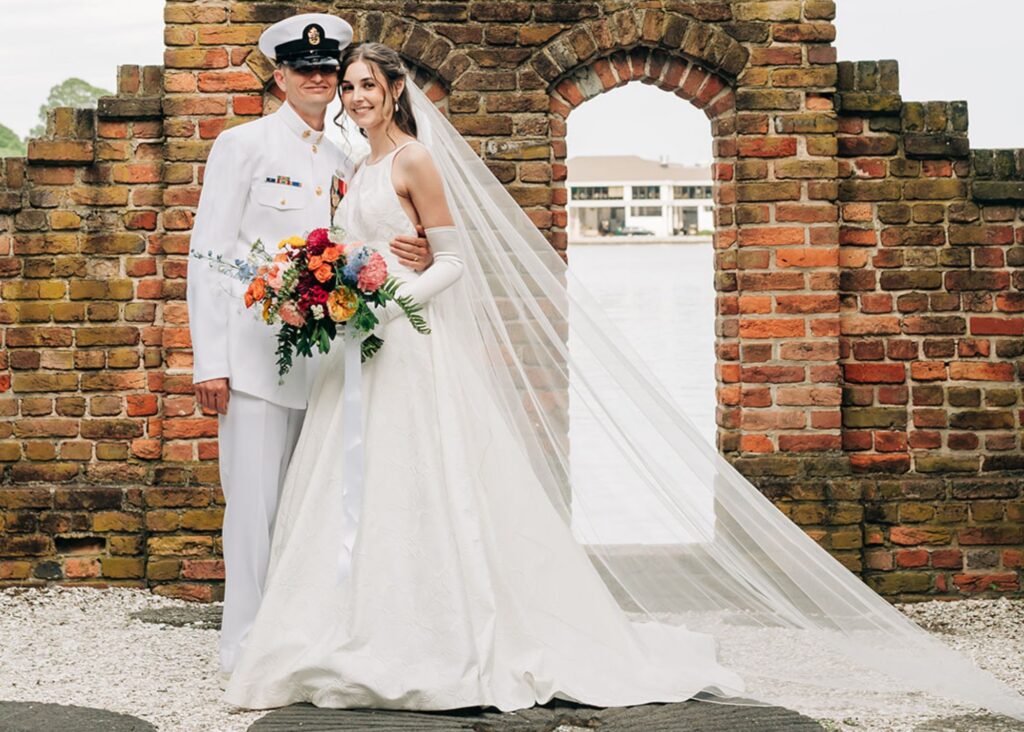 A brie and groom stand before a brick arch at the Hermitage Museum. The groom, a marine, wears his dress whites, the bride a jewel-necked ball gown wedding dress and long white gloves