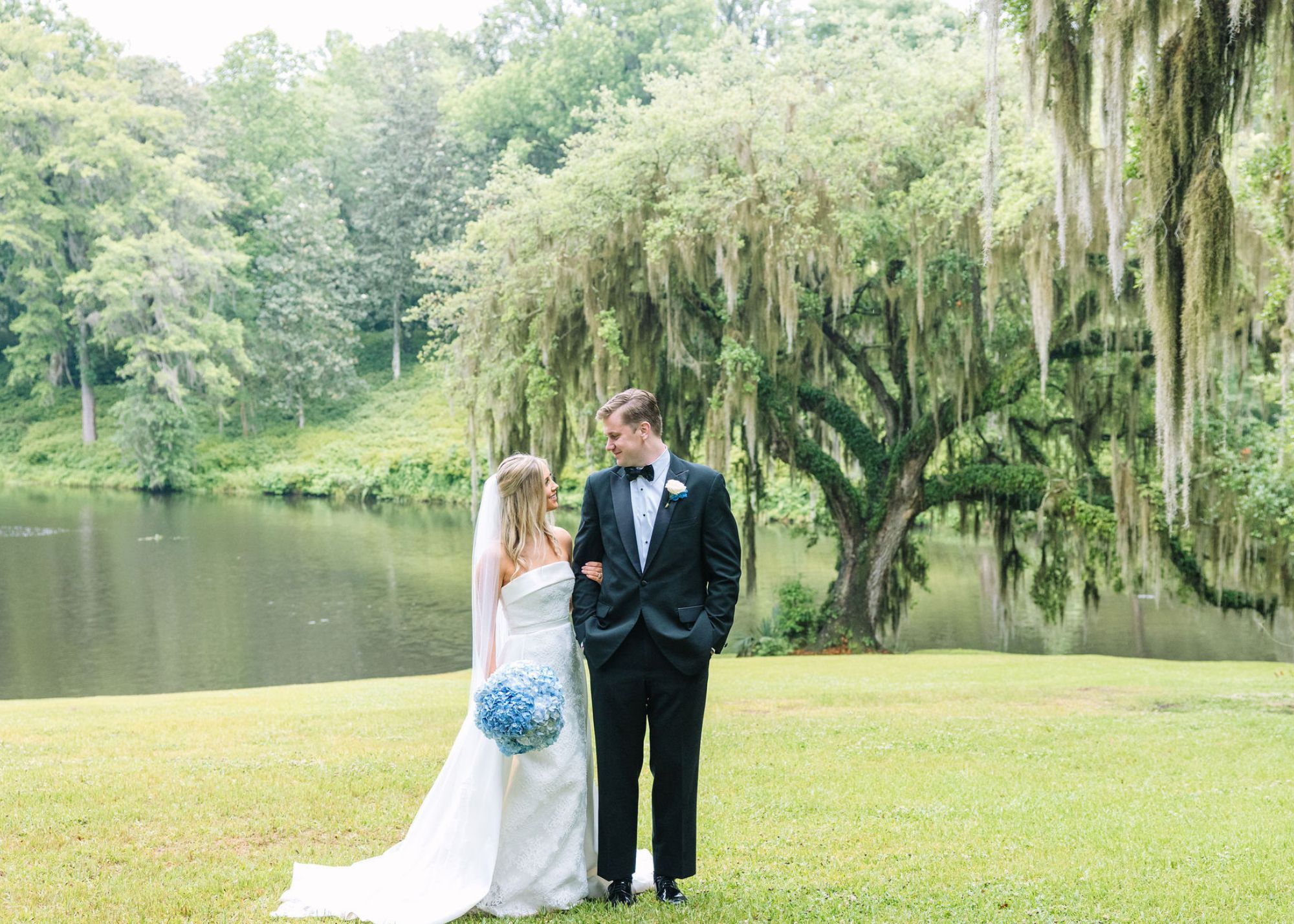 A bride and groom stand on a lawn with a spanish moss-draped oak in the background. The bride carries a blu hydrangea bouquet and wears her lace column gown and detachable overskirt by Lea-Ann Belter Bridal