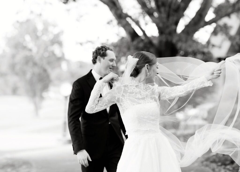 A black and white photo of a bride and groom - the bride's veil flies in the breeze, adding drama to the moment. The bride wears and all-lace ballgown wedding dress and sheer lace jacket with long sleeves.