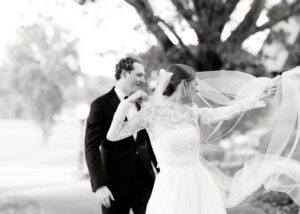 A black and white photo of a bride and groom - the bride's veil flies in the breeze, adding drama to the moment. The bride wears and all-lace ballgown wedding dress and sheer lace jacket with long sleeves.