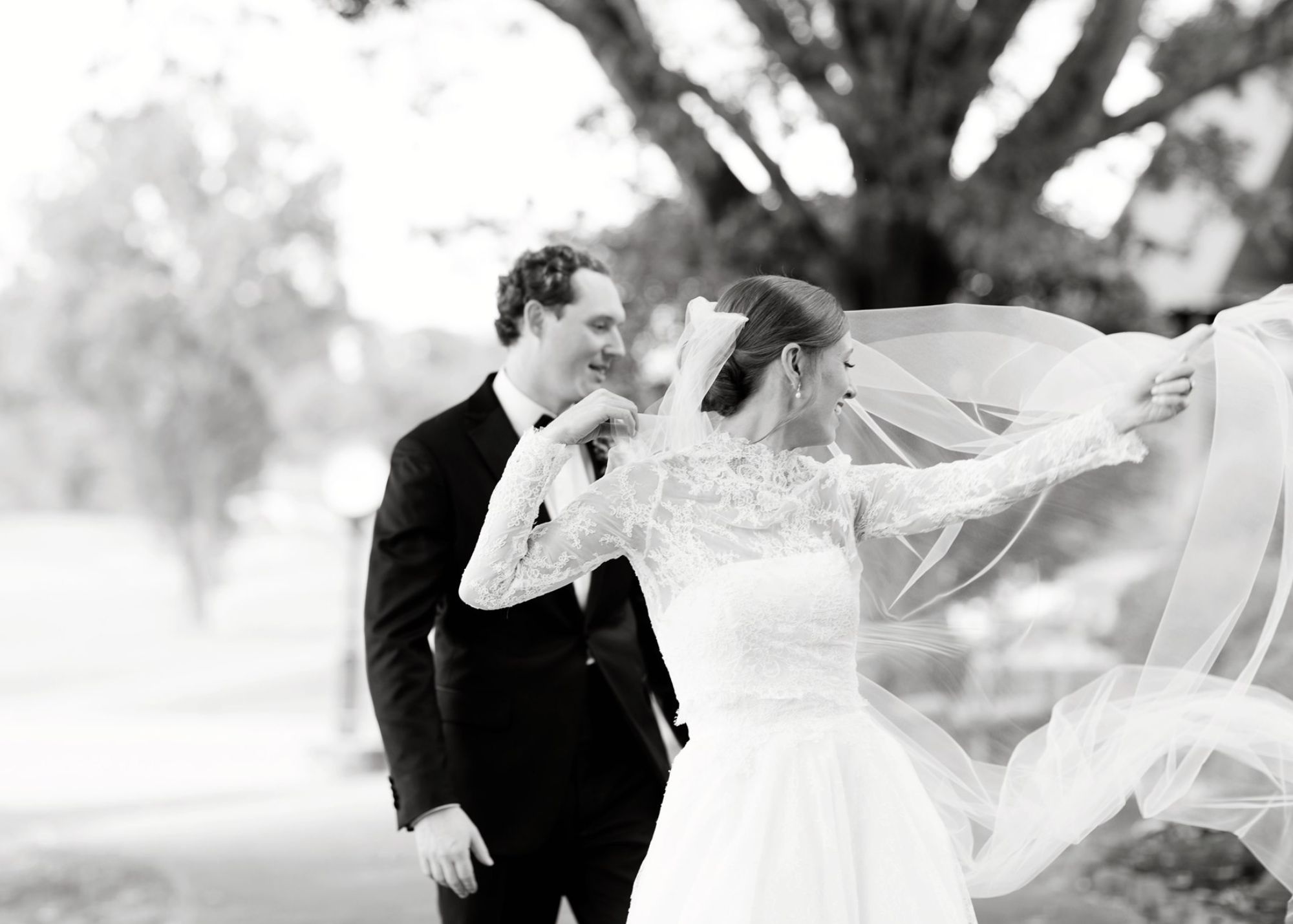 A black and white photo of a bride and groom - the bride's veil flies in the breeze, adding drama to the moment. The bride wears and all-lace ballgown wedding dress and sheer lace jacket with long sleeves.